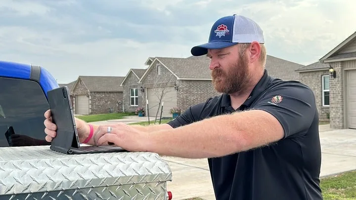 HVAC technician in Piedmont, OK, working on a tablet placed on a truck's diamond plate toolbox, with a residential neighborhood in the background, focused on the task at hand.