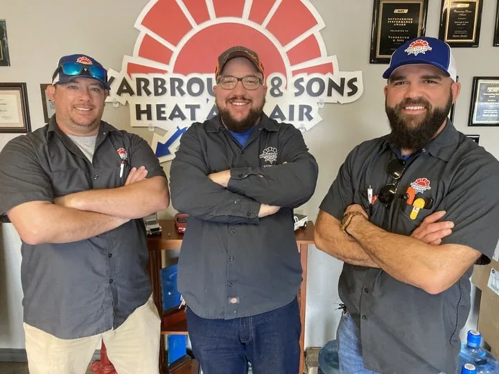 Three smiling HVAC technicians from Yarbrough & Sons Heat and Air standing confidently in front of the company logo, dressed in uniform shirts, with arms crossed, showcasing their team spirit and professionalism