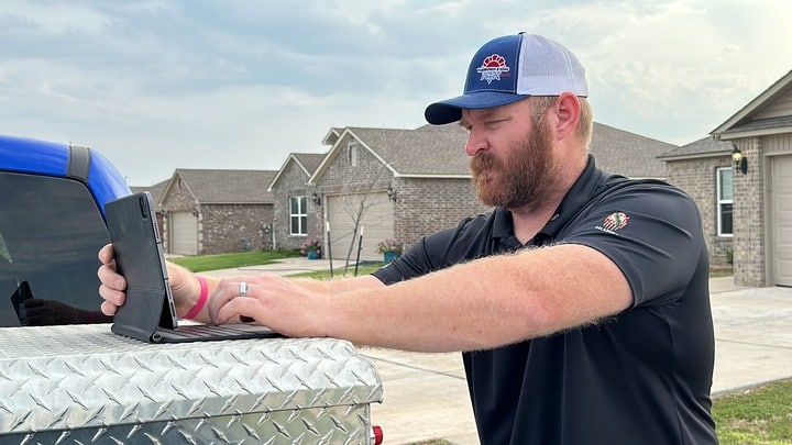 HVAC technician in Piedmont, OK, working on a tablet placed on a truck's diamond plate toolbox, with a residential neighborhood in the background, focused on the task at hand.