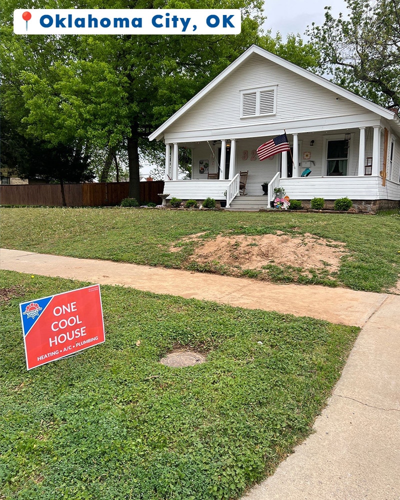 Yarbrough & Sons service yard sign in front of a white craftsman-style home in Oklahoma City, OK, with an American flag on the porch and fresh dirt from a recent plumbing repair visible in the yard.